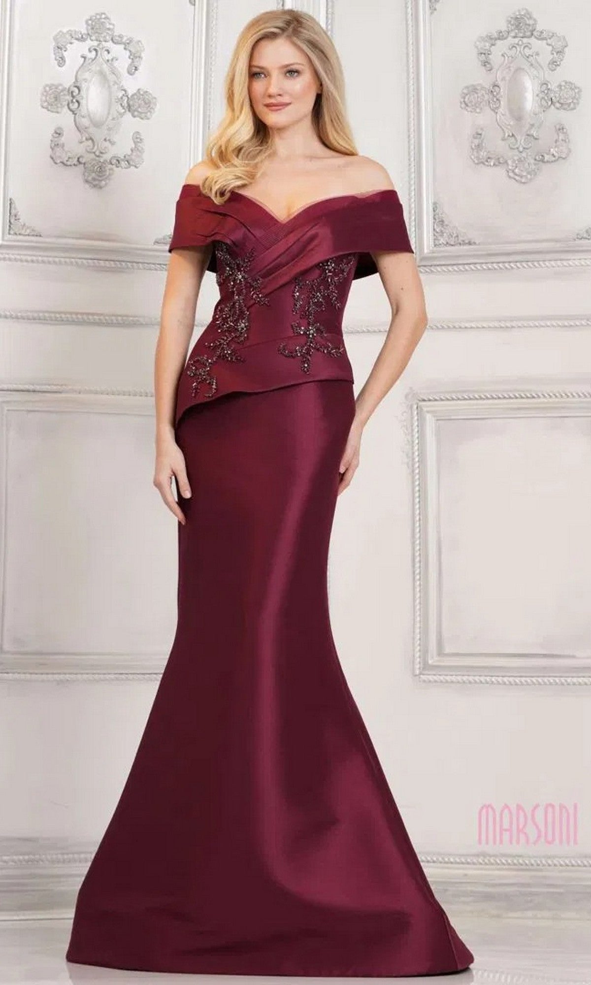A woman stands in front of a decorative white wall, wearing the Marsoni MV1312 long formal dress in burgundy—an off-shoulder gown with a mermaid skirt and floral bead embellishments.